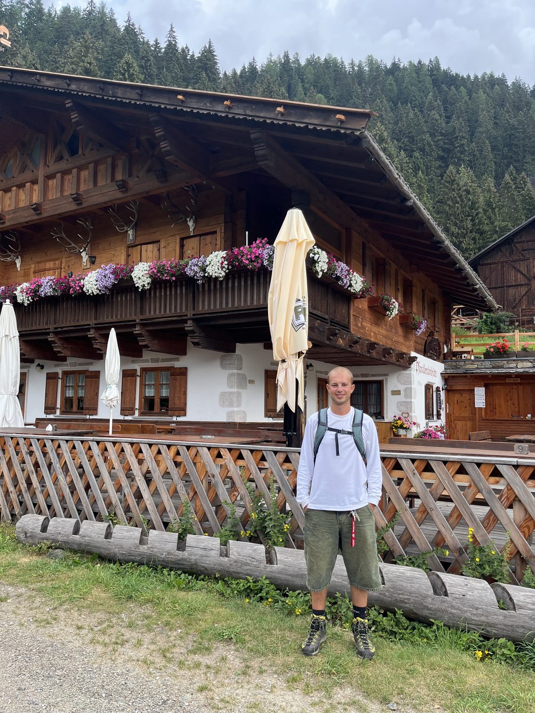 Alessio standing in front of a wooden Alpine farmhouse with flower boxes, forested slopes behind.