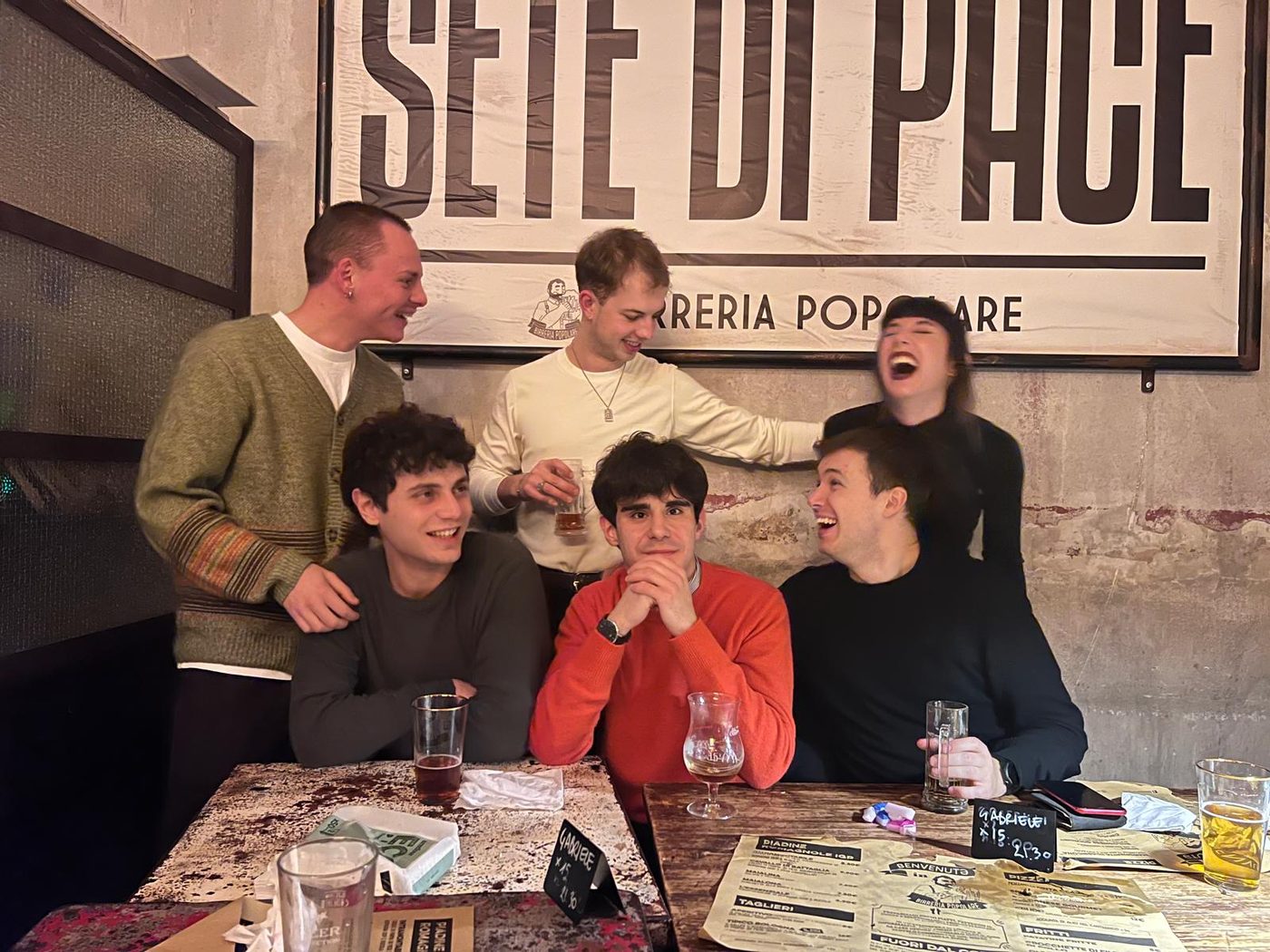 Six friends laughing around a table in a bar, under a large sign reading Sete di Pace.
