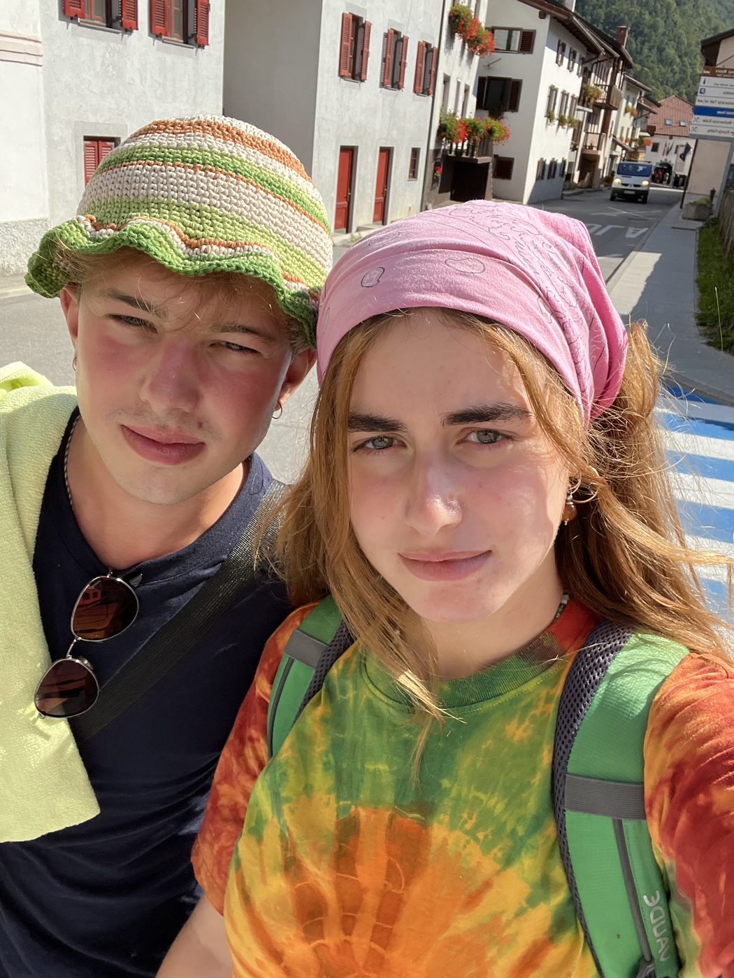 Alessio in a crocheted bucket hat and a friend in a pink headscarf and tie-dye shirt, both carrying backpacks, on the street of an Alpine village.